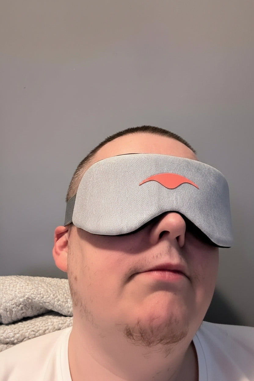 Person wearing a gray sleep mask with a brand logo on a plain background